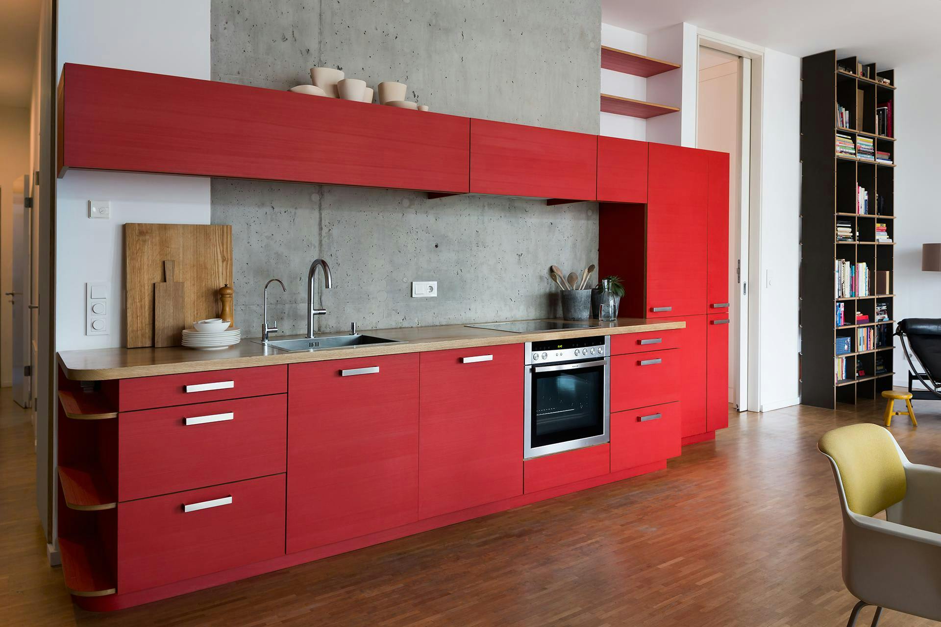 The image showcases a modern, red kitchen with a large island and a stainless steel oven. The kitchen is well-equipped with various appliances, including a microwave, a toaster, and a sink. The island is surrounded by a bookshelf, which is filled with numerous books.

The kitchen also features a dining table with a chair placed nearby. The room is decorated with a vase, a bowl, and a cup, adding a touch of elegance to the space. The overall atmosphere of the kitchen is bright and inviting, making it an ideal spot for
