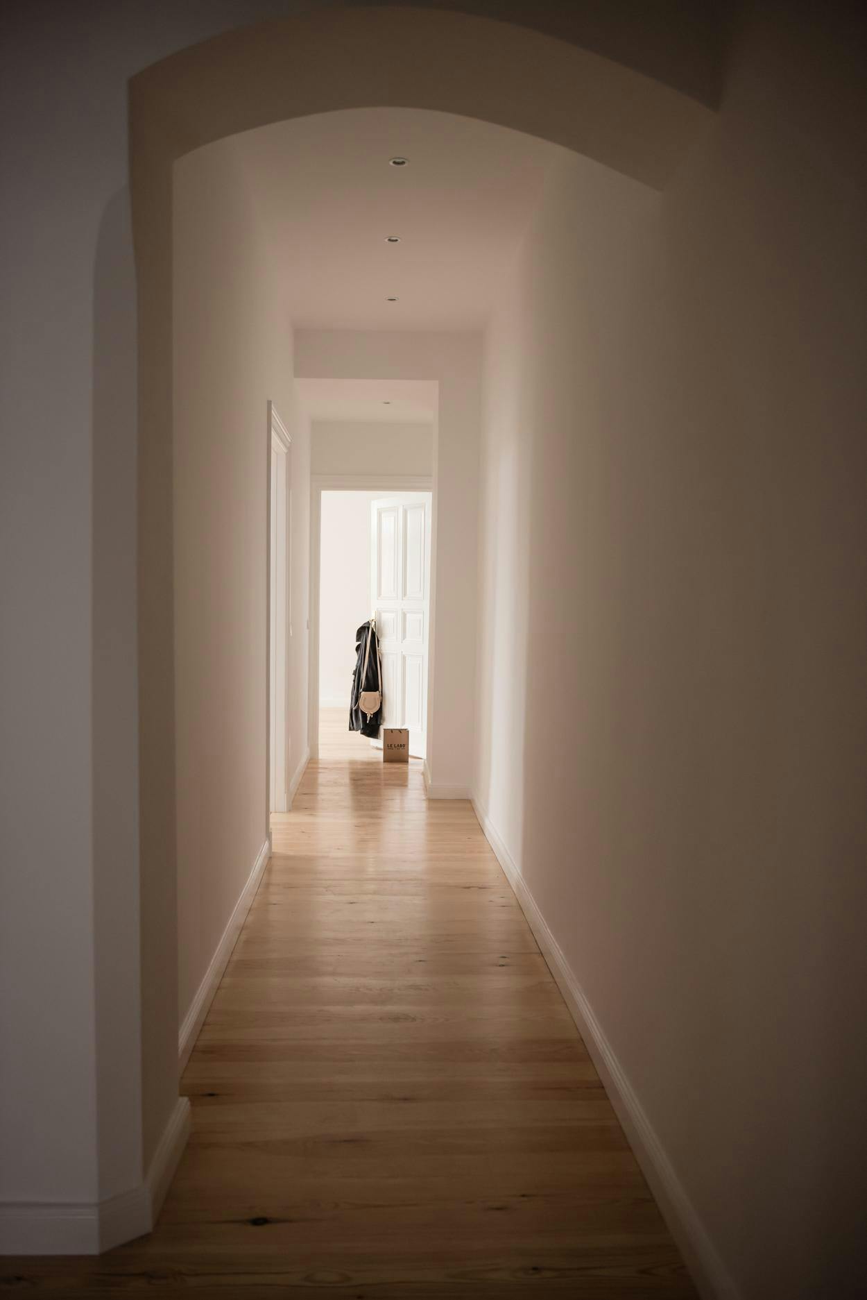 The image shows a long, narrow hallway with a wooden floor. The hallway is empty, with no people or objects visible. The lighting in the hallway is dim, creating a somewhat eerie atmosphere. The wooden floor extends from one end of the hallway to the other, and the walls are painted white. The hallway appears to be empty and quiet, making it a suitable setting for a movie or a quiet moment.