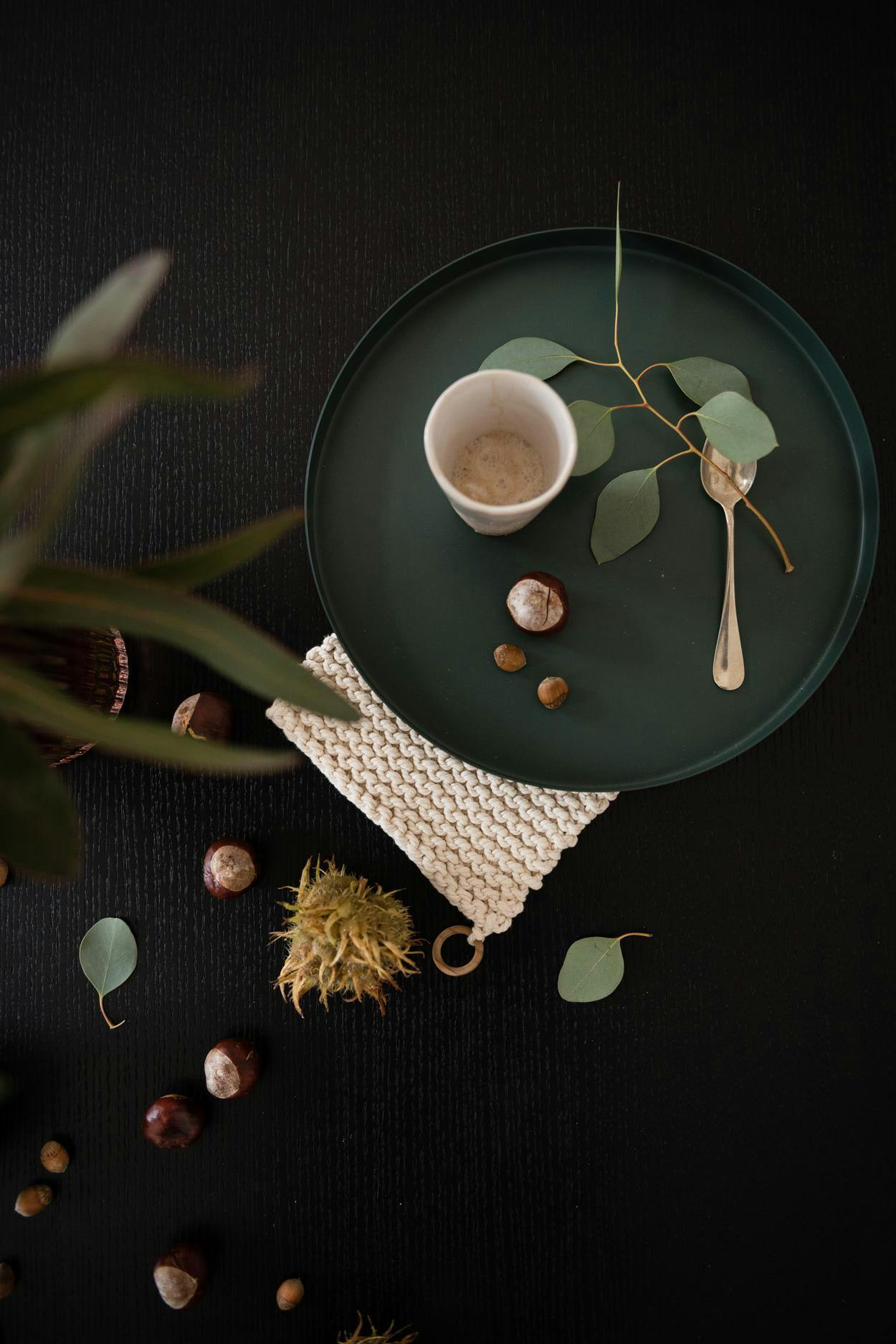 The image features a green plate with a cup of coffee and a spoon on it. The plate is placed on a table, and the coffee is poured into the cup. There are also some small pine cones scattered around the plate, adding a touch of nature to the scene.

In addition to the main plate, there are two smaller pine cones placed on the table, one near the left edge and the other near the right edge. These small pine cones add a decorative element to the scene, further enhancing the overall aesthetic.