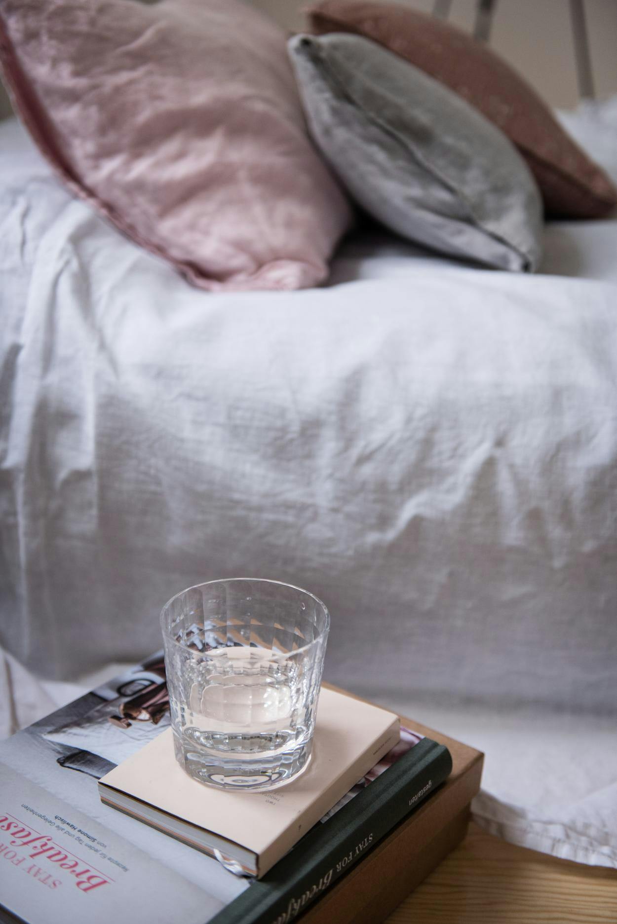 The image features a bed with a white comforter and a glass of water on top of it. The glass is placed on a small table, which is also on the bed. The bed appears to be a twin bed, and the glass of water is positioned near the edge of the bed.

In addition to the glass of water, there is a book on the bed, which is placed on the left side of the bed. The book appears to be a small, thin book, possibly a travel guide or a guide to a specific location. The scene suggests a cozy and relaxing atmosphere, with the b