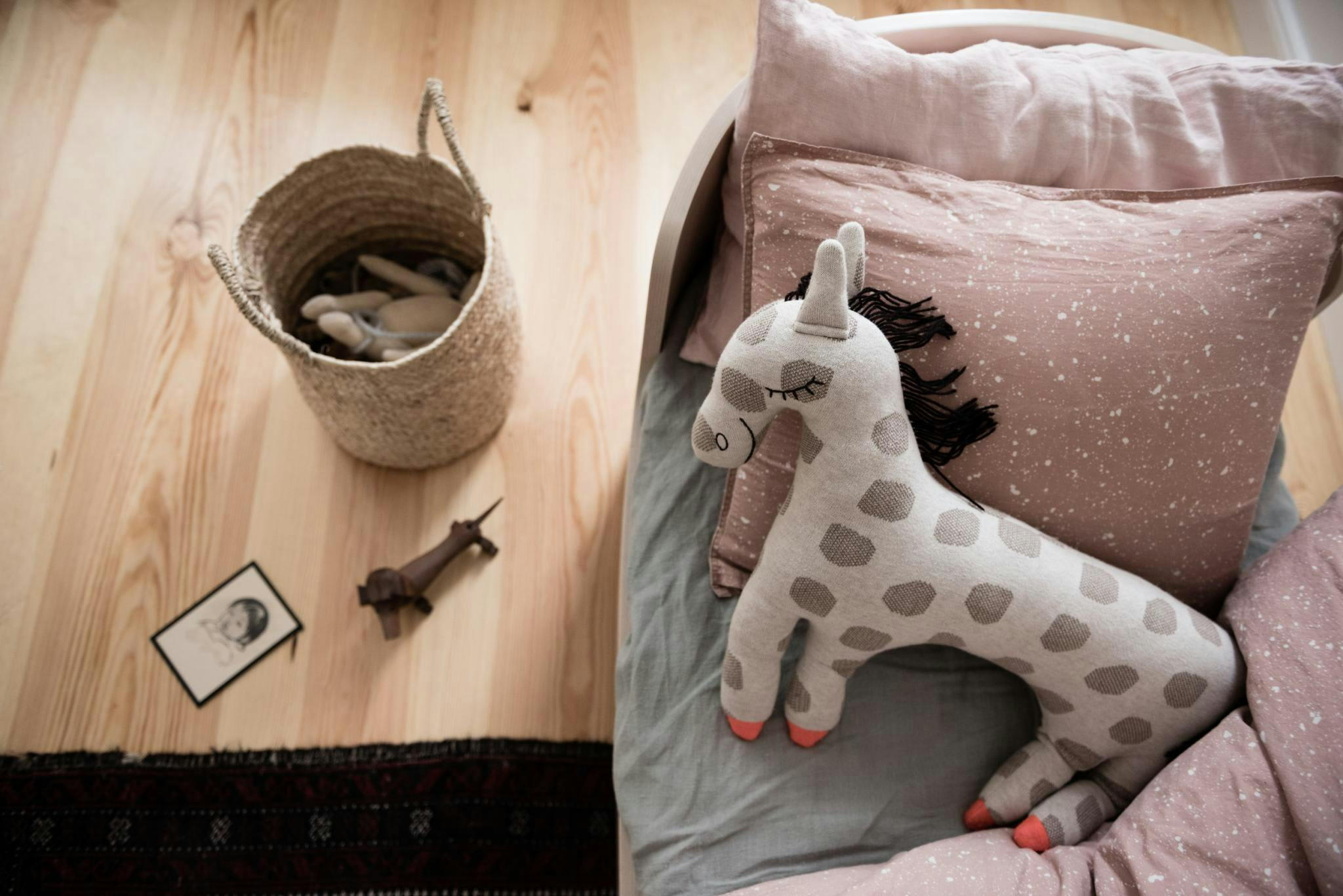 The image features a bedroom with a wooden floor and a bed. On the bed, there is a small stuffed horse, which is a popular toy for children. The horse is placed next to a basket, which is also on the bed.

In addition to the bed and the stuffed horse, there are two books on the bed, one on the left side and the other on the right side. A clock is also visible on the wall, adding to the cozy atmosphere of the room.