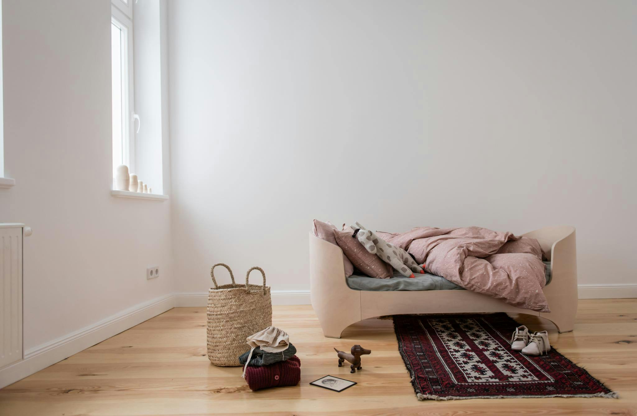 The image features a small, white couch in a room with a wooden floor. The couch is placed in the center of the room, and a basket is placed next to it. There are two handbags on the floor, one near the couch and the other slightly further away. 

In addition to the couch and handbags, there are two books on the floor, one closer to the couch and the other slightly further away. A remote control is also visible on the floor, likely for controlling the television or other electronic devices in the room.