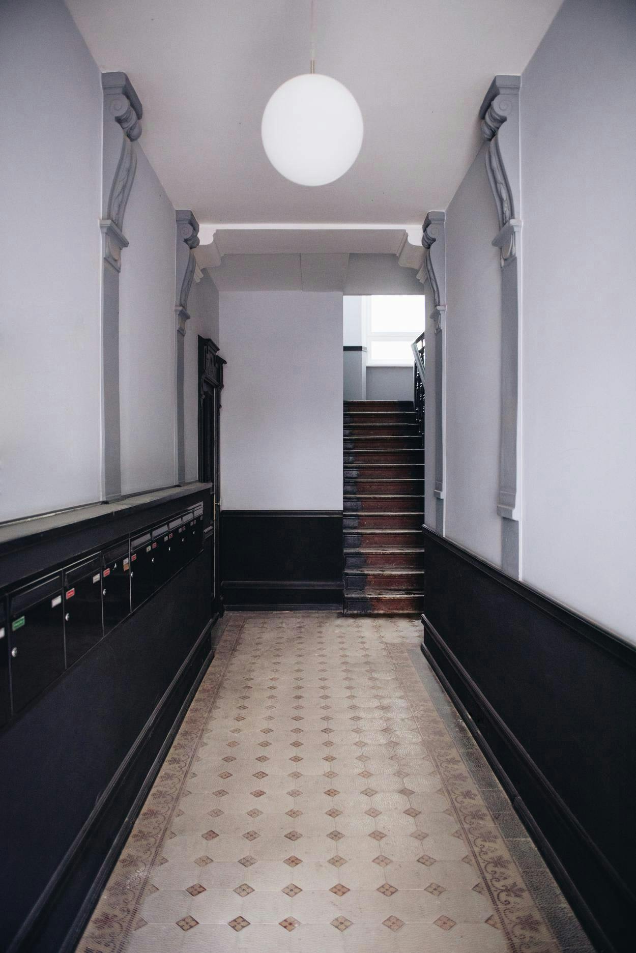 The image shows a long, narrow hallway with a black and white checkered floor. The hallway is empty, with no people or objects visible. The hallway is lit by a large, round, white light fixture, which is situated above the stairs. The light fixture is positioned at the top of the stairs, providing a bright and welcoming atmosphere. The hallway appears to be empty and clean, making it a suitable space for a quiet walk or a peaceful retreat.