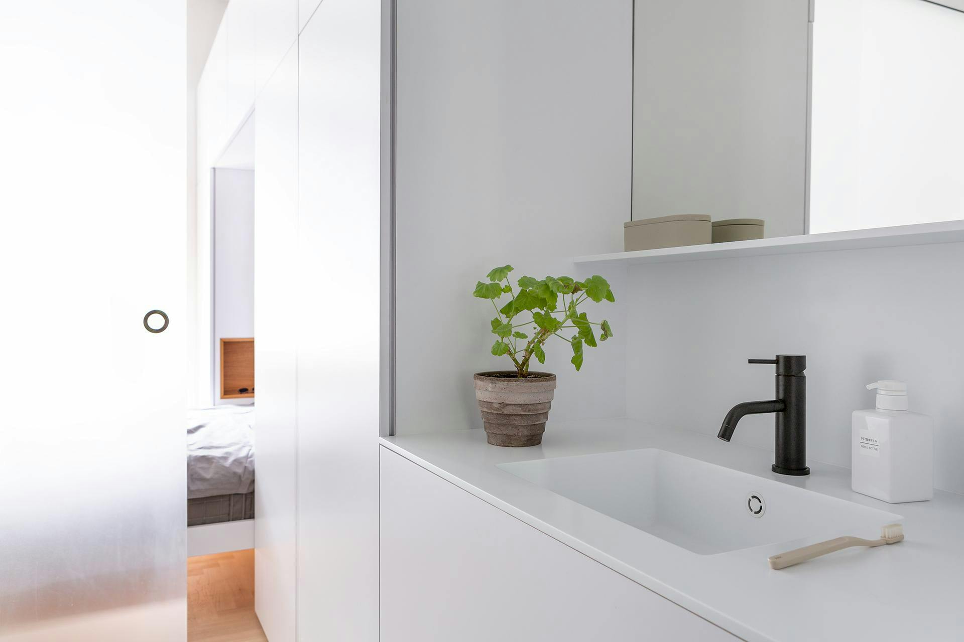 The image features a clean, white bathroom with a sink and a mirror. The sink is located on the left side of the room, while the mirror is positioned above it. A potted plant is placed on the right side of the room, adding a touch of greenery to the space.

In addition to the sink and mirror, there are two bottles in the bathroom. One bottle is located on the left side of the room, while the other is situated on the right side, closer to the potted plant. There is also a toothbrush placed near the sink, indicating that the bath