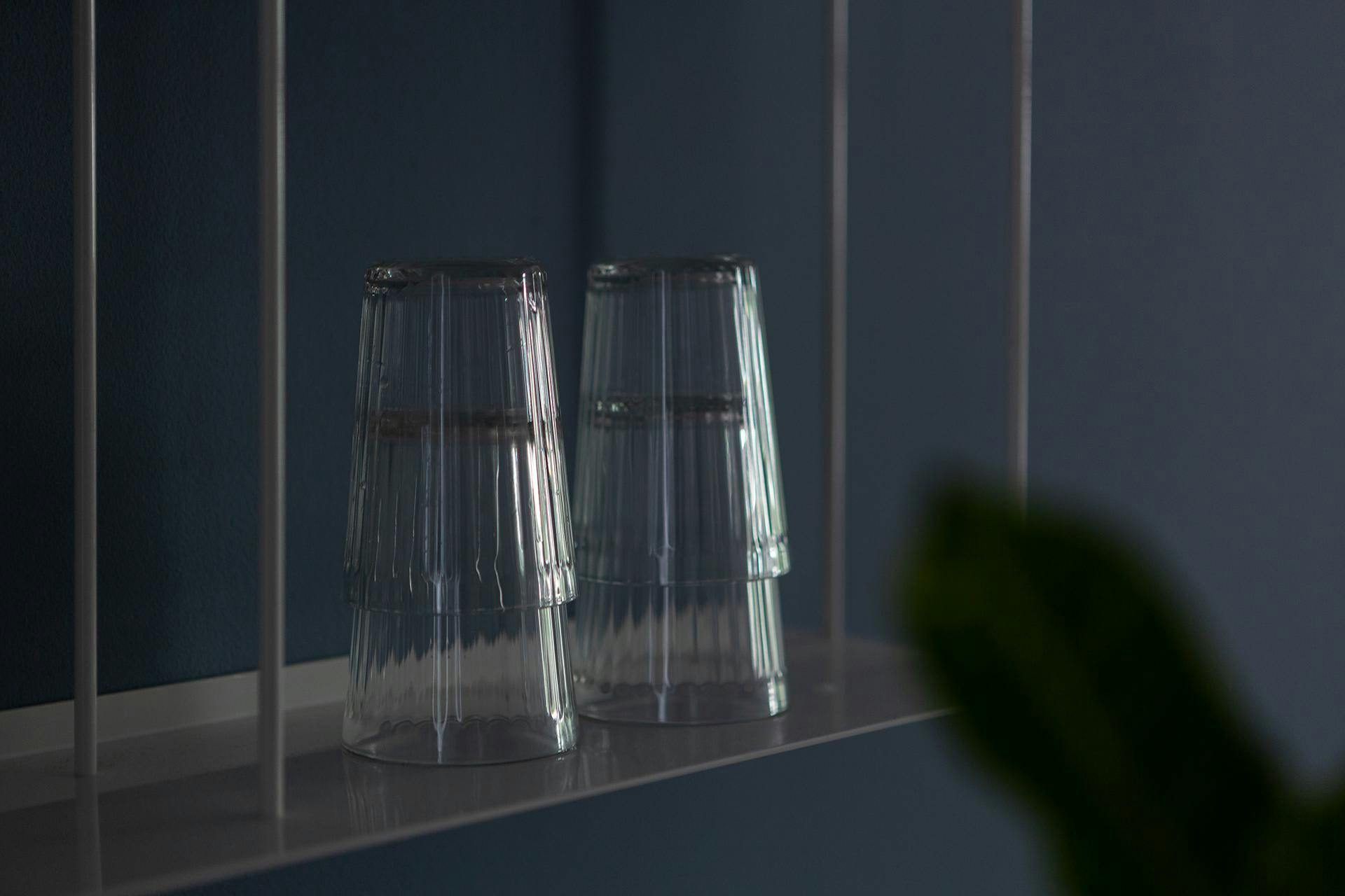 The image features a white shelf with a row of clear glass vases or jars lined up against the wall. There are four vases in total, each with a unique shape and size. The vases are arranged in a neat and organized manner, creating an aesthetically pleasing display. The clear glass vases are placed close together, adding a sense of harmony and balance to the arrangement.