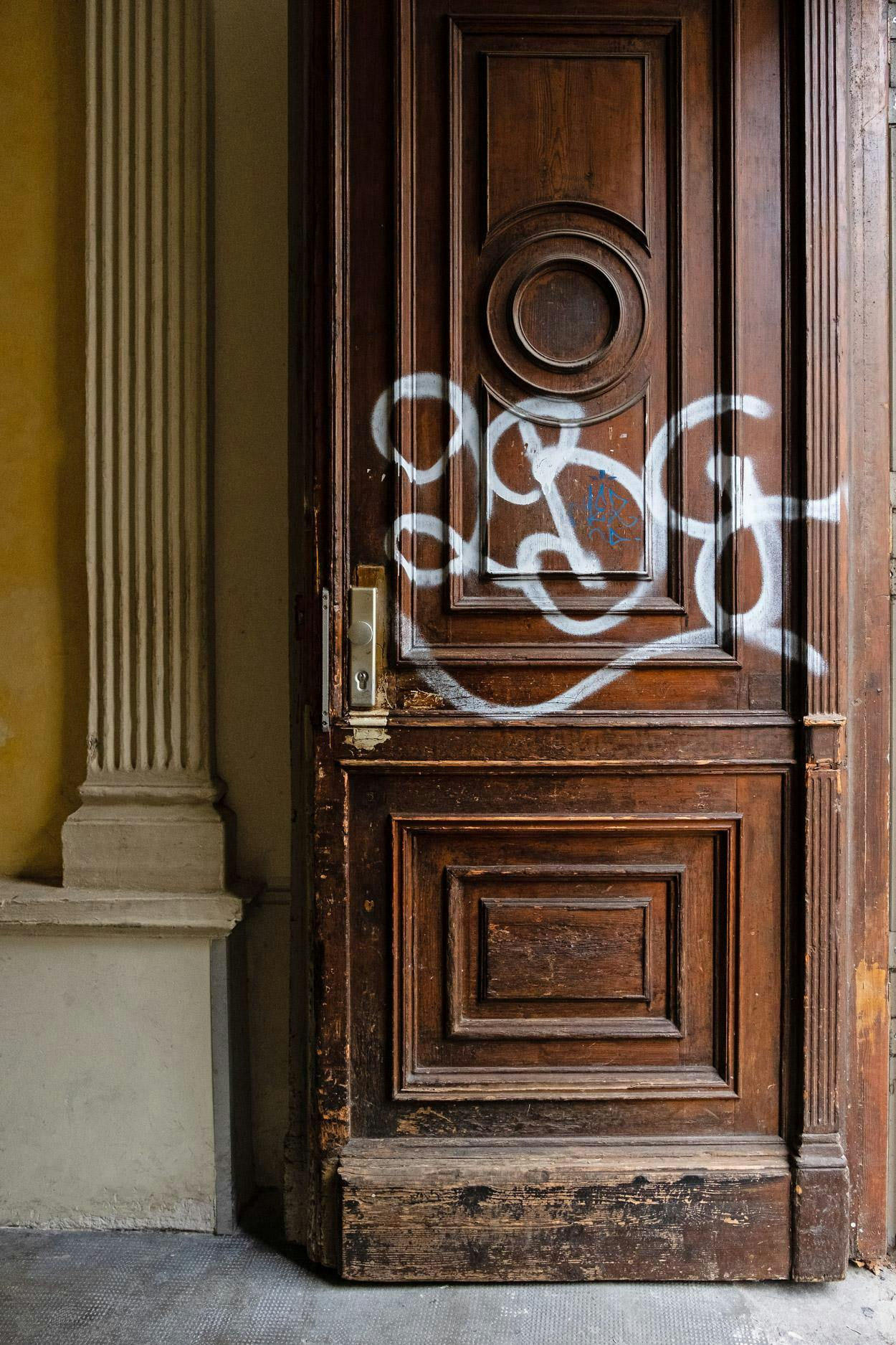 The image features a large wooden door with a graffiti-covered "G" on it, which is open and located next to a building.