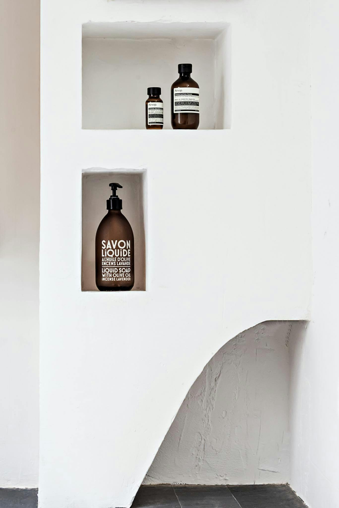 A white shelf with a bottle of soap and a bottle of shampoo is displayed in a bathroom setting.