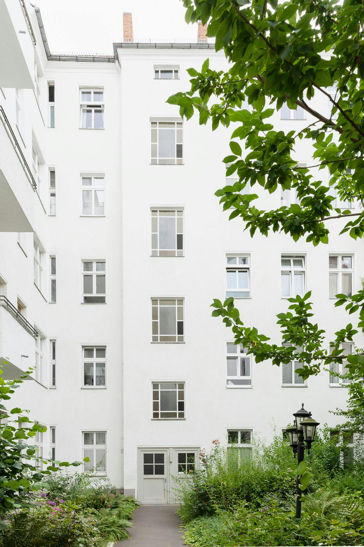 A large white building with many windows is situated next to a lush green garden, with a tree in front of it.