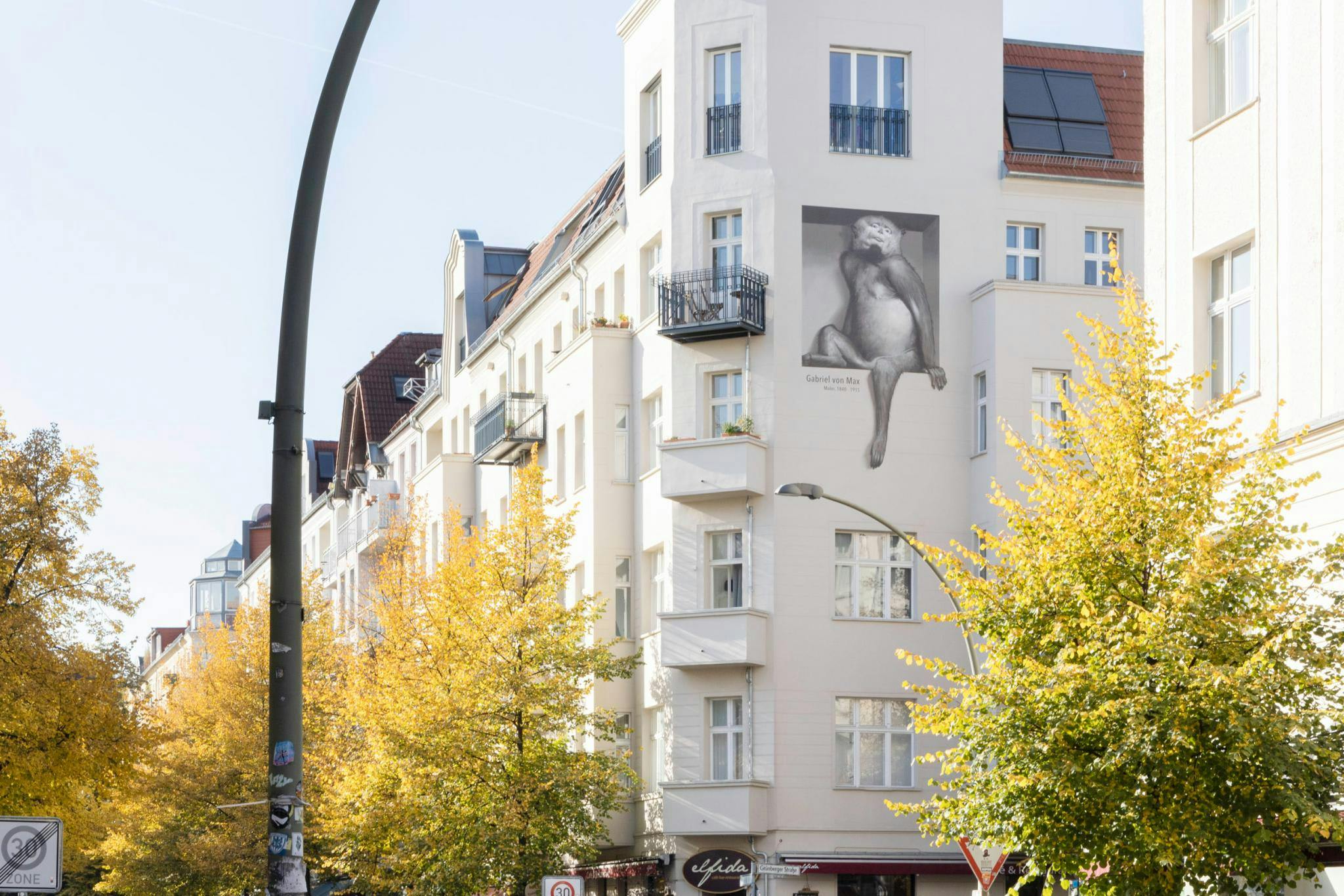 A large white building with a mural of a woman on its side is located in a city, surrounded by trees and a street.