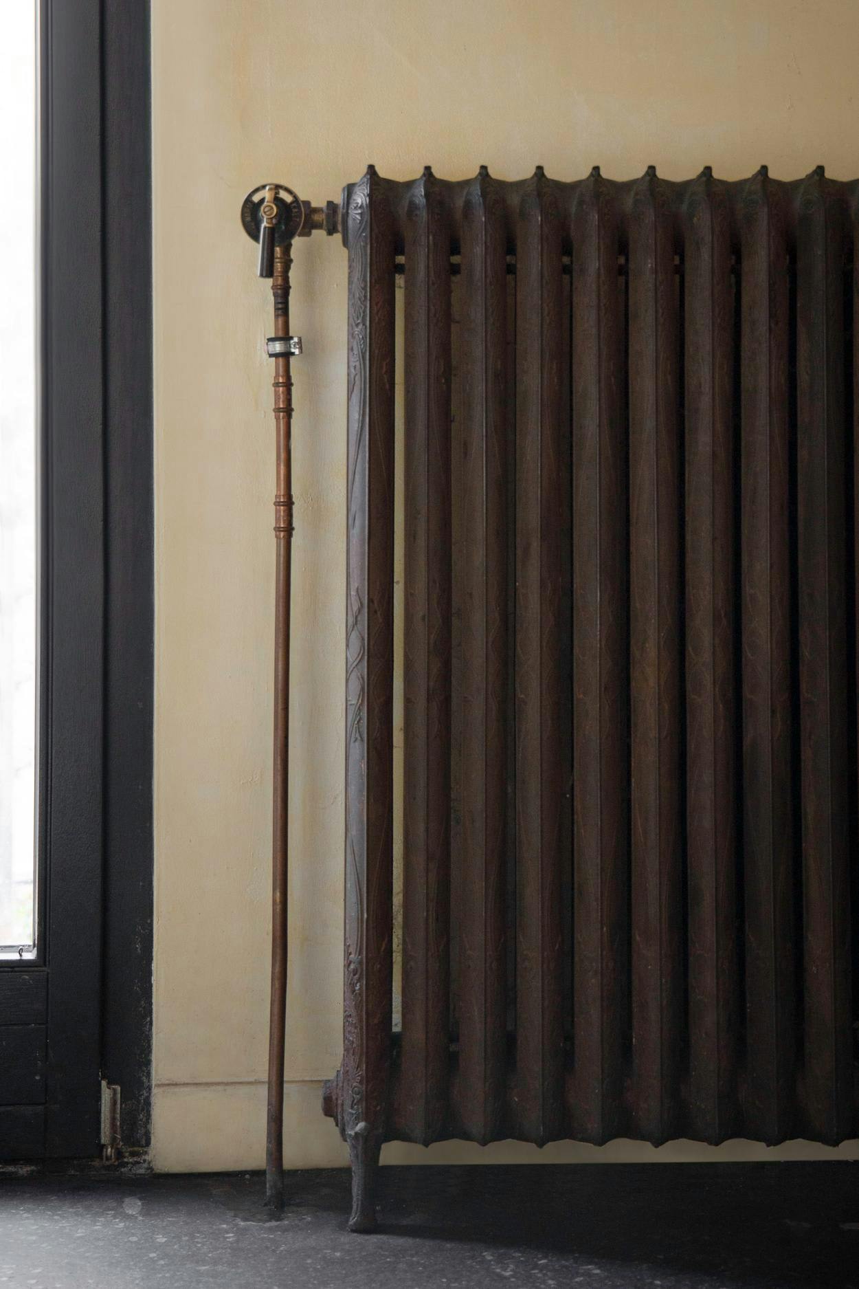 A large, old-fashioned radiator is mounted on a wall next to a window, with a metal pipe or pipe cover attached to it.