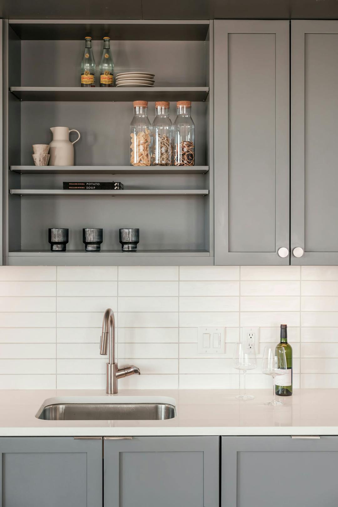 a kitchen with gray cabinets and a stainless steel sink and a wine bottle on a shelf above the sink